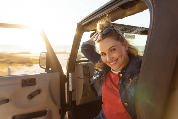 一个白人女人坐在敞着的顶层轿车里 开著车门 面带微笑地对着镜头 眼前的景象正在逼近她 — 图库照片