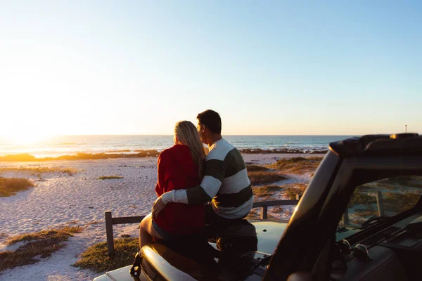 一对白种人夫妇在敞篷轿车外的后视镜 背景是夕阳西下的海滩 他们拥抱在一起 望着远方 — 图库照片