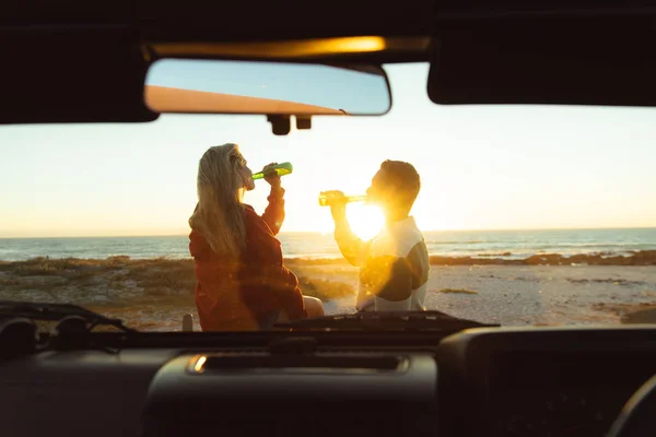 一对白种人夫妇坐在敞篷轿车外的后视镜 背景是夕阳西下的海滩 喝啤酒 — 图库照片