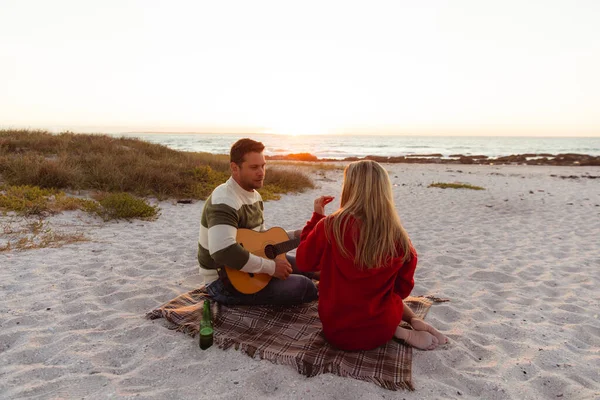一对白种人夫妇斜倚在海滩上的背影 背景是夕阳 男人拿着吉他 女人拿着食物 — 图库照片