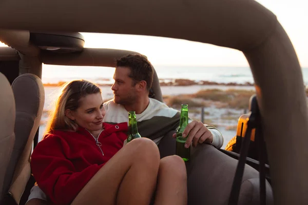 オープントップカー内の白人カップルのフロントビュー バックグラウンドでビーチで日没と ビールのボトルを包含し — ストック写真