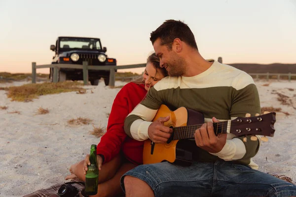 一对白种人夫妇斜倚在海滩上 后面是一辆敞篷轿车 男人拿着吉他 女人抱着她的伴侣 拿着一瓶啤酒 — 图库照片