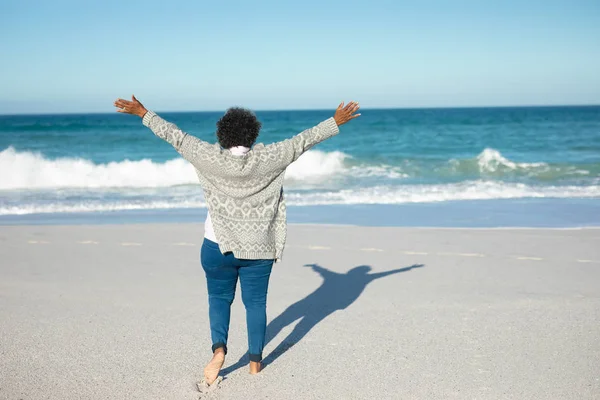 一位非洲裔美国老年妇女站在海滩上的后视镜 背景是蓝天和大海 她举起胳膊望着地平线 — 图库照片