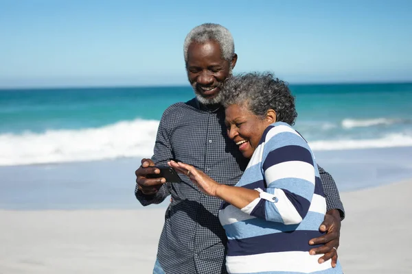 青い空と海に囲まれたビーチに立つアフリカ系アメリカ人の高齢者のカップルの側面図 抱擁と彼らの携帯電話を使用して — ストック写真