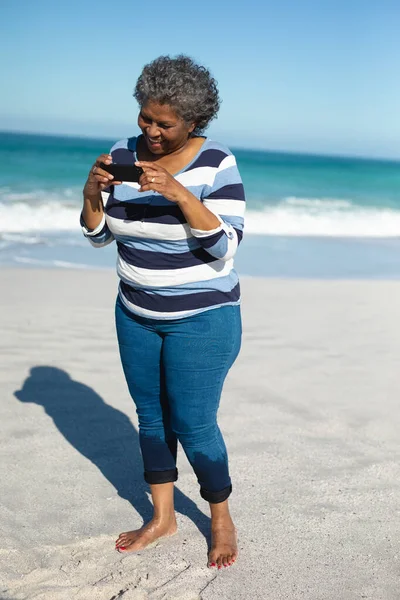 一位站在海滩上 赤着脚 蓝蓝的天空和大海为背景 微笑着 拿着她的手机的非洲裔美国女人的前景 — 图库照片