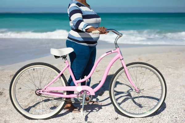 一位非洲裔美国妇女站在海滩上的中间 背景是蓝天和大海 她骑着粉色的自行车 — 图库照片
