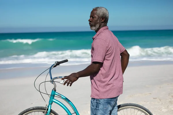 青い空と海を背景に青い自転車を持って遠くを見ているアフリカ系アメリカ人の老人の正面図 — ストック写真