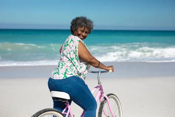 一位非洲裔美国老年妇女骑自行车在海滩上的镜头 背景是蓝天蓝海 转身对着镜头微笑 — 图库照片