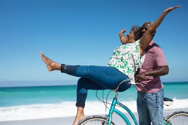 在海滩上 一对背景为蓝天和大海的非洲裔美国老夫妻的侧面照片 当这位女士坐在扶手上的时候 这位男士把她推上了自行车 — 图库照片