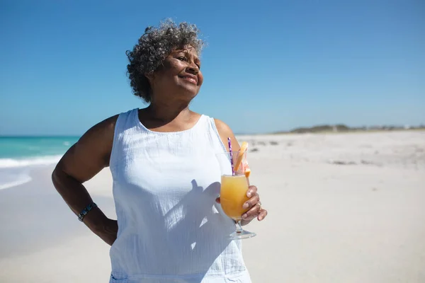 一位非洲裔美国老妇人站在沙滩上 站在阳光下 举着鸡尾酒 面带微笑 背景是蓝天和平静的大海 — 图库照片