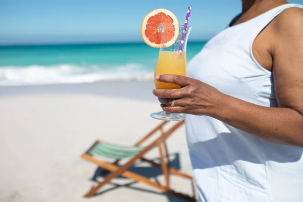 一个女人站在海滩上站在阳光下 拿着鸡尾酒 在海滩上有一张躺椅 背景是蓝天和平静的大海 — 图库照片