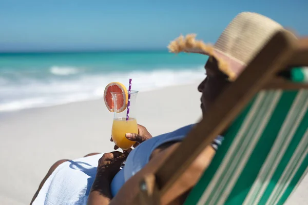 デッキチェアに座って帽子をかぶってカクテルを飲みながら青い空と穏やかな海を背景にしたアフリカ系アメリカ人のシニア女性の側面図 — ストック写真