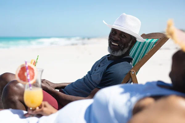 一对非洲裔美国老夫妻坐在阳光下的海滩上 坐在甲板上 戴着太阳帽 拿着鸡尾酒 转过身来 面带微笑 背景是蓝天和平静的大海 — 图库照片
