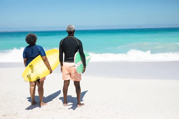 青い空と背景に穏やかな海と 彼らの腕の下にサーフボードを保持し 太陽の下でビーチでシニアアフリカ系アメリカ人カップルの背面ビュー — ストック写真