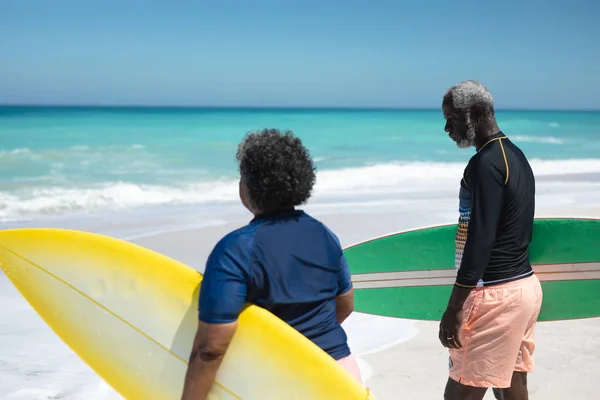青い空と背景に穏やかな海と 彼らの腕の下にサーフボードを保持し 太陽の下でビーチで上級アフリカ系アメリカ人のカップルの側面図 — ストック写真