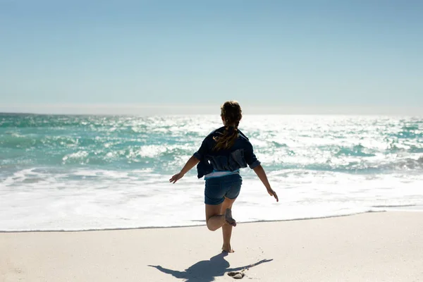 青い空と海を背景に 腕を伸ばして走る日当たりの良いビーチで白人の少女の背面図 — ストック写真