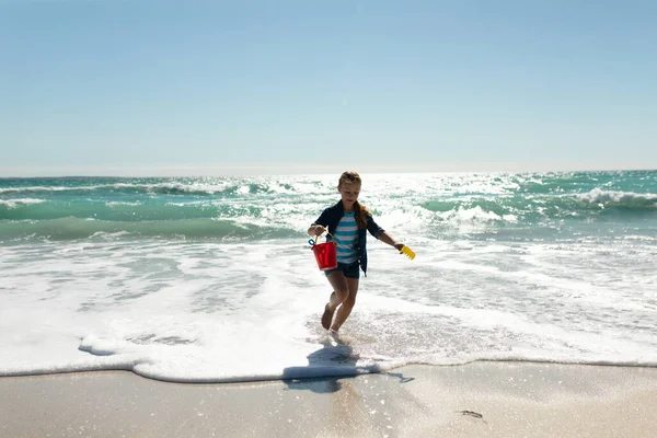 青い空と海を背景に バケツとスペードを保持カメラに向かって砂の上に裸足で実行され 日当たりの良いビーチで白人の女の子のフロントビュー — ストック写真