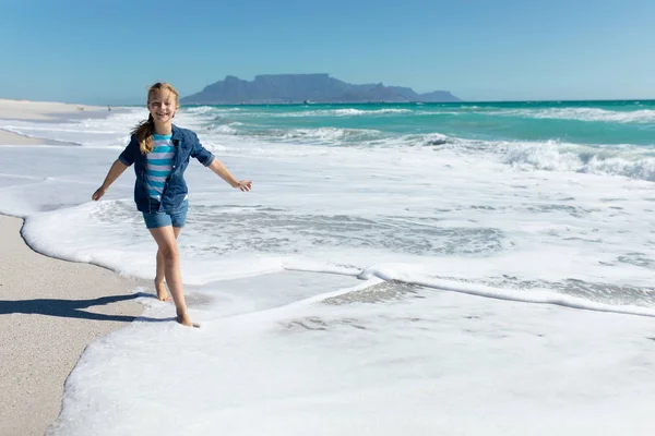 一个白人女孩站在阳光普照的海滩上 走在浅浅的水面上 微笑着对着镜头 背景是蓝天和大海 — 图库照片