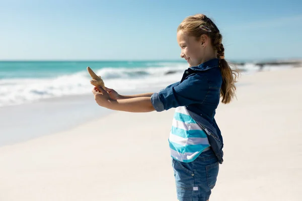 一个白人女孩站在阳光灿烂的海滩上 手里拿着一只海星 望着镜头 微笑着 背景是蓝蓝的天空和大海 — 图库照片