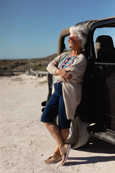 太陽の下でビーチでのシニア白人女性の側面図は 腕を組んで車にもたれて交差サングラスを着用し 離れて笑みを浮かべて見て — ストック写真