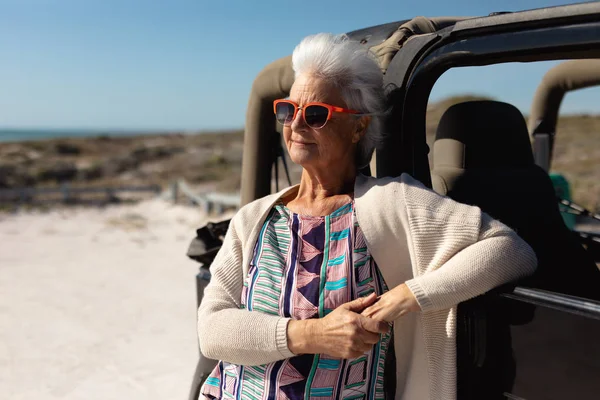 車にもたれかかったりサングラスをかけたり笑顔を見送ったりしながら太陽の下のビーチで白人の先輩女性の正面図 — ストック写真