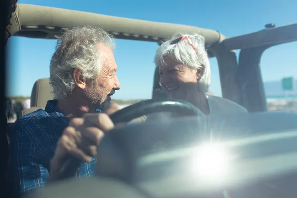 車の中で 太陽の下でビーチに運転して座っているシニア白人のカップルのフロントビューでは ホイールで男は お互いを見て 笑みを浮かべて フロントガラス レンズフレアを介して見られる — ストック写真