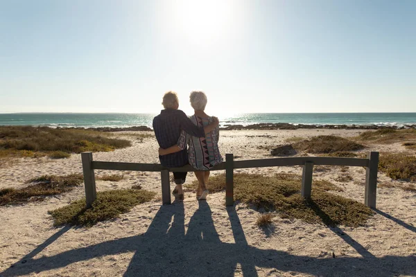 一对年长的白种人夫妇坐在沙滩上晒太阳 坐在栅栏上 欣赏海景 拥抱在一起的背景图 — 图库照片