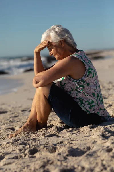 太陽の下でビーチでは シニア白人女性の側のビューは 砂の上に座って 背景に青い空で彼女の目をシールド — ストック写真