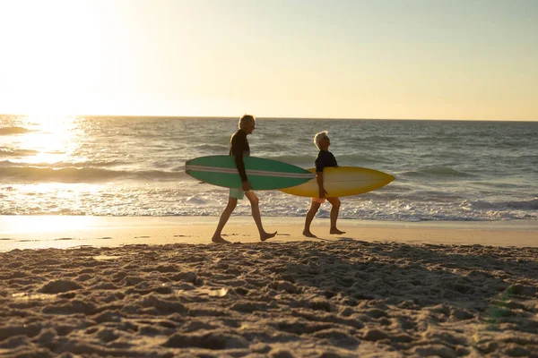 夕阳西下 一对年长的白种人夫妇抱着冲浪板在海滩上散步时的侧影 — 图库照片