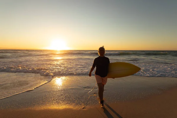 夕日のビーチで白人の先輩女性の正面ビュー 彼女の腕の下にサーフボードを運ぶカメラに向かって砂の上を歩いて — ストック写真
