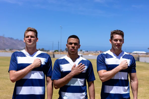 Front view of two Caucasian male rugby players and a mixed race male rugby player, all wearing a team strip, standing on a pitch with their right hands over their hearts listening to their national anthem