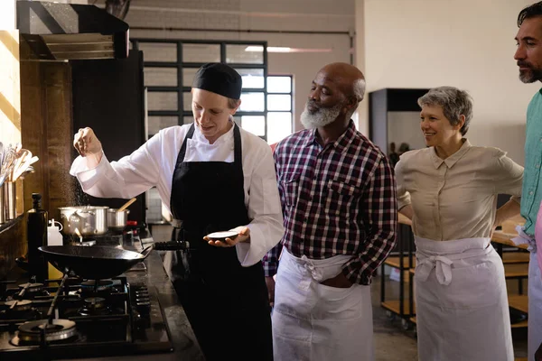 stock image Side view of multi-ethnic Senior group at cookery class, discussing, listening to a teacher, who is pouring salt at a frying pan