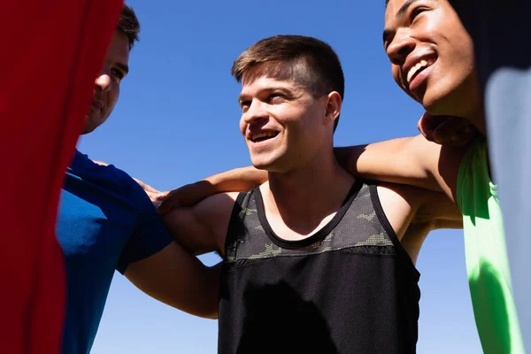 青い空に対して太陽の下で笑顔肩の周りの腕を持つスポーツフィールドでの男性ランナーのトレーニングの多民族グループの低角度フロントビュー — ストック写真