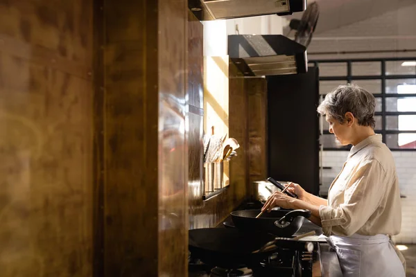 レストランのキッチンでの調理教室での白人シニア女性の側面図 窓の前で鍋で調理し スマートフォンを使用して — ストック写真