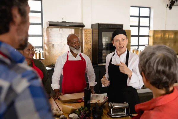 調理師の授業では 多民族の高齢者の肩の上に 白人の料理人と黒の帽子とエプロンを着た白人女性シェフの指示に耳を傾ける多様な大人の学生 — ストック写真
