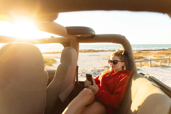 一个白人女人坐在敞着的顶层汽车里 用她的手机 面带微笑 周末海滩度假 生活方式和休闲 — 图库照片