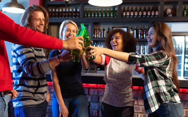 Side view of a group of five multi-ethnic male and female friends, standing at the bar in a pub, holding bottles of beer and raising them to make a toast, and laughing together