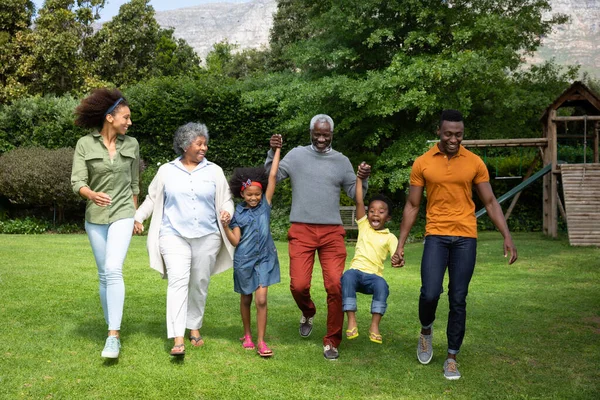 Front view of a multi-generation African American family in the garden, walking together, all holding hands, the grandfather and father lifting the grandson in the air and smiling.