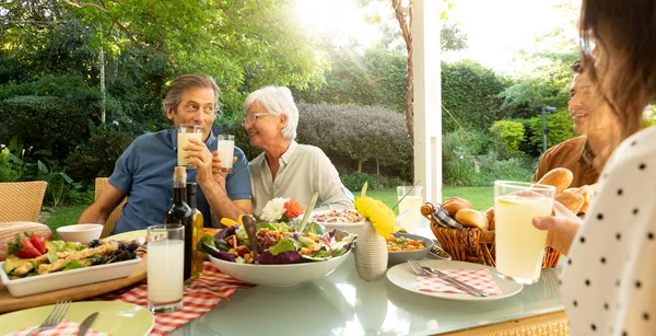 一对年长的白种人夫妇和家人坐在外面的餐桌旁吃饭 两臂挽着 面带微笑地喝酒 他们的成年儿子和他年幼的女儿站在了前面 — 图库照片