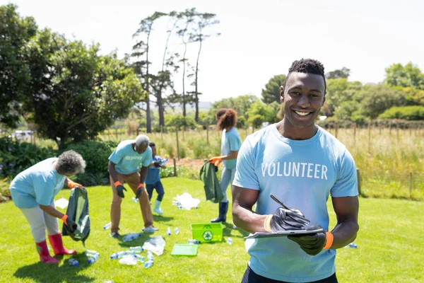 Tシャツを着たアフリカ系アメリカ人の笑顔が描かれたフィールドに ボランティアが手袋を着用し クリップボードに書き込み カメラに笑顔で立っている姿が描かれ さまざまなボランティアグループがゴミやリサイクルを収集しています — ストック写真