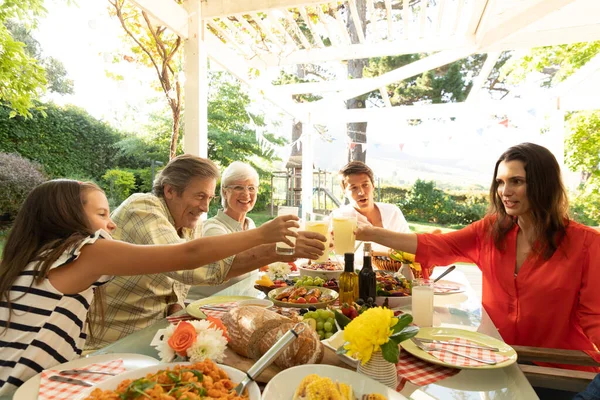 複数世代の白人の家族の正面ビューは 食事のためのディナーテーブルセットの外に座って 一緒にメガネを上げ トーストを作る — ストック写真