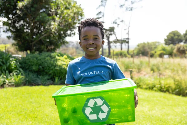 ボランティアが書かれたTシャツを着たフィールドに立つアフリカ系アメリカ人の少年の笑顔を正面から眺め 緑のプラスチックのリサイクル箱を持ち カメラに微笑む — ストック写真