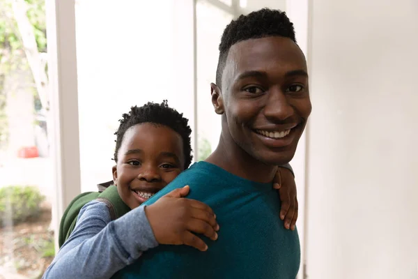 幸せなアフリカ系アメリカ人の男の家で彼の息子のピギーバックを運ぶとカメラに笑顔の肖像画 — ストック写真