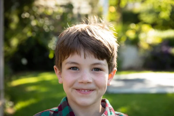 若い白人の男の子の肖像画で 短い茶色の髪と青い目がカメラに微笑む日当たりの良い庭の外に立っています — ストック写真