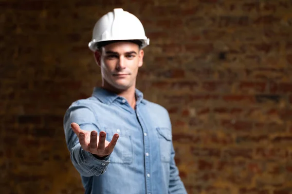 一个白种人建筑工程师的画像 他黑发短发 头戴白色头盔 头戴斜纹棉布衬衫 他的手在面前触摸着一个以砖墙为背景的虚拟互动屏幕 — 图库照片