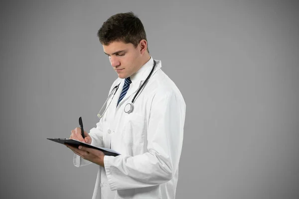 聴診器付きのラボコートを身に着けている白人男性医師のサイドビュー 灰色の背景にクリップボードのメモを保持立って — ストック写真