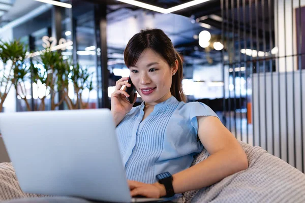 現代のオフィスで働くスマートな服を着たアジア人実業家の女性が 豆の袋に座ってノートパソコンやスマートフォンを使って — ストック写真