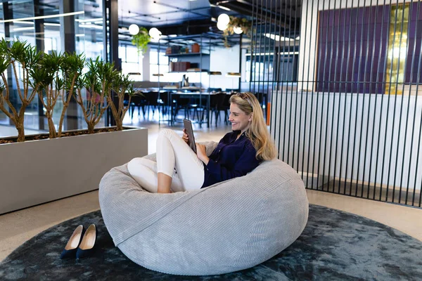 現代のオフィスで働くスマートな服を着た白人実業家の正面図 ビーンバッグに座ってタブレットを使用する — ストック写真