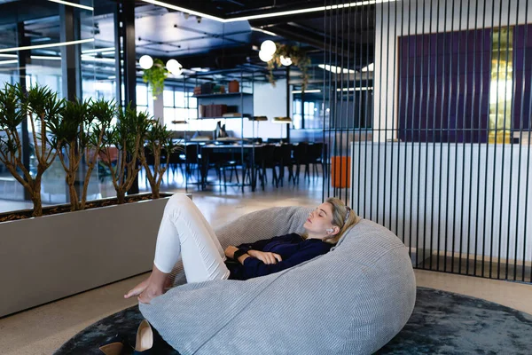 一个身穿漂亮衣服和耳机的白人女商人 在现代化的办公室工作时休息一下 躺在豆袋上休息的侧影 — 图库照片