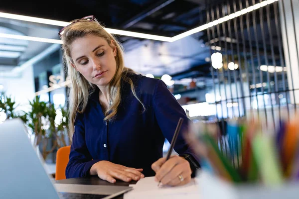 現代のオフィスで仕事をし机のそばに座り紙に書いている白人実業家の女性の正面図 — ストック写真